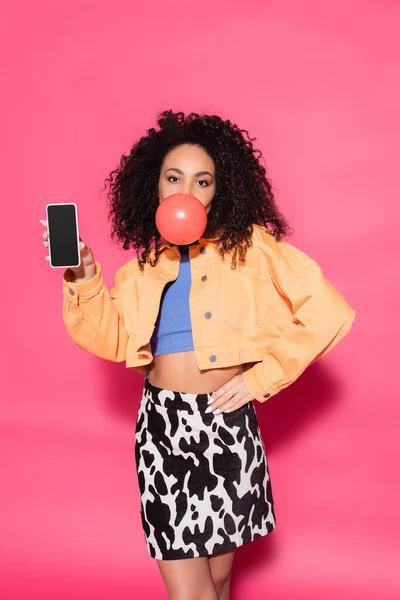 Encaracolado Africano americano mulher soprando chiclete e segurando smartphone com tela em branco no rosa — Fotografia de Stock