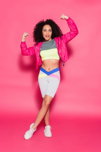 Full length of sporty brunette african american woman showing muscles while posing on pink — Stock Photo