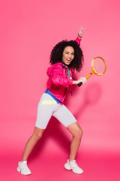 Piena lunghezza di donna africana americana stupita e sportiva che tiene la racchetta da tennis sul rosa — Foto stock