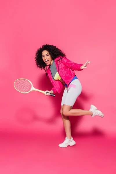 Pleine longueur de femme afro-américaine excitée tenant raquette de tennis sur rose — Photo de stock