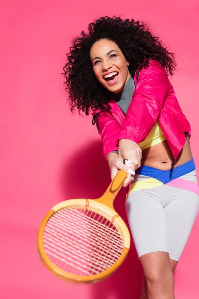 Mujer afroamericana asombrada sosteniendo raqueta y jugando tenis en rosa - foto de stock