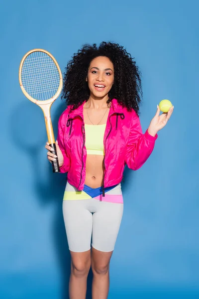Mujer afroamericana emocionada sosteniendo raqueta de tenis y pelota en azul - foto de stock