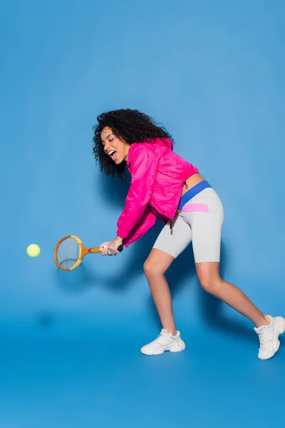 Longitud completa de la mujer afroamericana emocionada jugando tenis en azul - foto de stock
