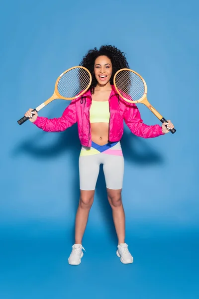 Full length of excited african american woman holding tennis rackets on blue — Stock Photo