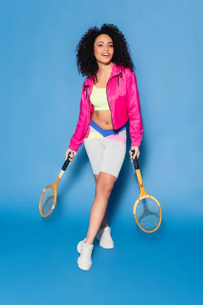 Piena lunghezza di allegra giovane donna afroamericana che tiene racchette da tennis su blu — Foto stock