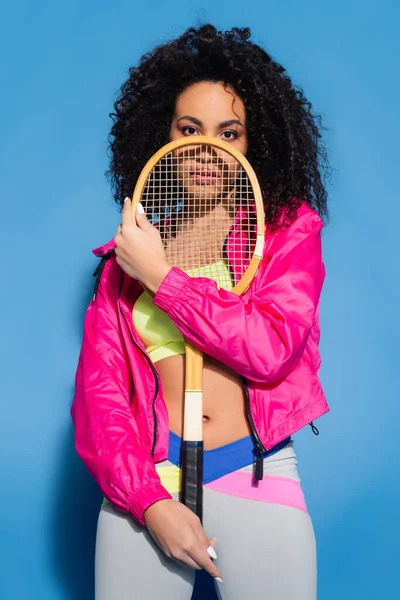 Encaracolado jovem afro-americana posando com raquete de tênis e olhando para a câmera em azul — Fotografia de Stock