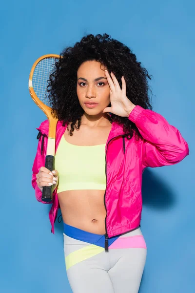 Rizado africano americano mujer en crop top posando con raqueta de tenis y mirando a la cámara en azul - foto de stock