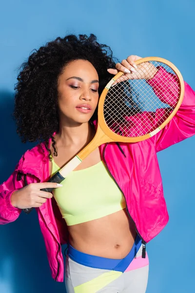 Curly african american woman in crop top posing with tennis racket on blue — Stock Photo