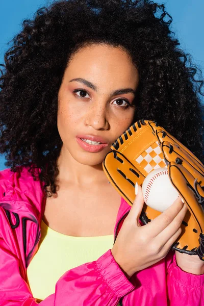 Lockige afrikanisch-amerikanische Frau in Lederhandschuhen hält Baseball isoliert auf blau — Stockfoto
