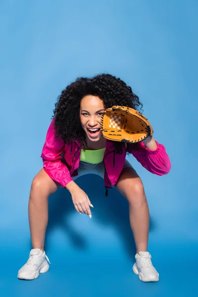 Full length of excited african american woman in leather glove playing baseball on blue — Stock Photo
