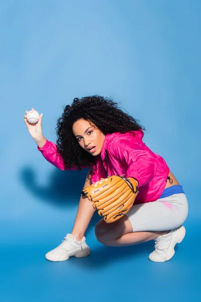 Lockige afrikanisch-amerikanische Frau in Lederhandschuhen wirft Baseball auf blau — Stockfoto