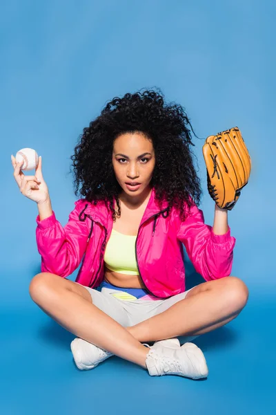 Curly african american woman in leather glove holding baseball while sitting with crossed legs on blue — Stock Photo