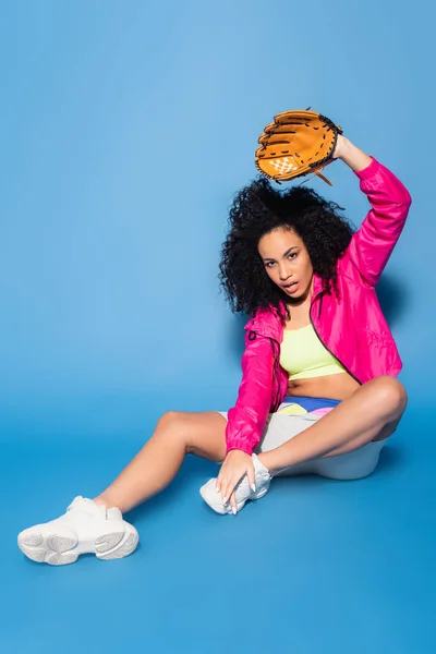 Mujer americana africana rizada en guante de béisbol de cuero sentado con la mano levantada en azul - foto de stock