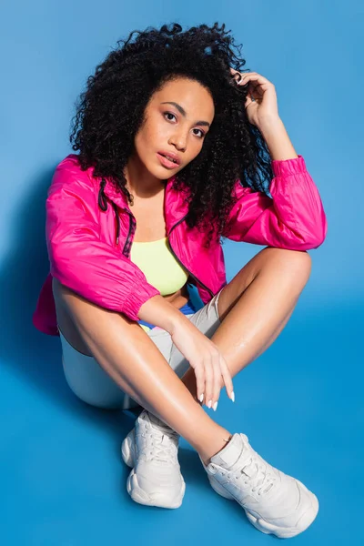 Femme afro-américaine bouclée en veste rose assise sur bleu — Photo de stock