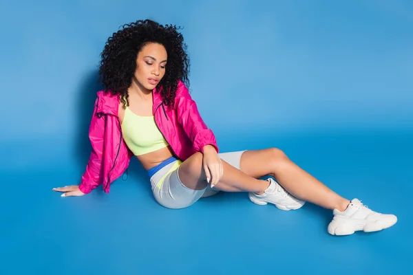 Full length of curly african american woman in pink jacket sitting on blue — Stock Photo