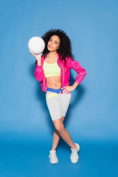 Full length of sporty african american woman in pink jacket holding volleyball while posing with hand on hip on blue — Stock Photo