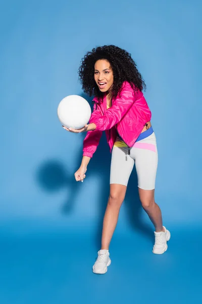 Piena lunghezza di donna afroamericana stupita in giacca rosa giocare a pallavolo su blu — Foto stock
