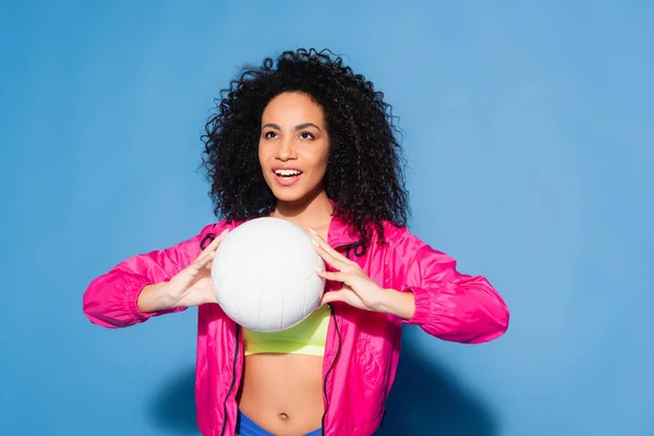 Happy african american woman in pink jacket and crop top playing volleyball on blue — Stock Photo