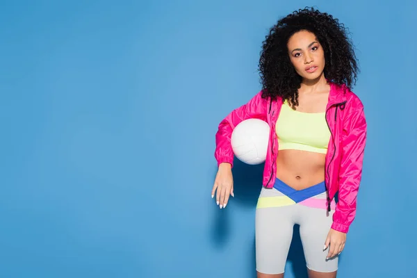 Curly african american woman in pink jacket and crop top standing with volleyball on blue — Stock Photo