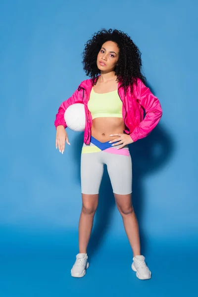 Full length of curly african american woman in pink jacket and crop top posing with volleyball on blue — Stock Photo