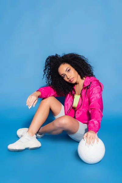 Young african american woman in pink jacket sitting near volleyball on blue — Stock Photo