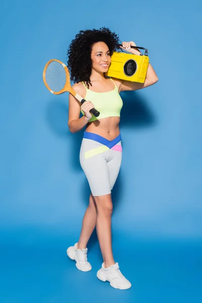 Comprimento total de mulher americana africana feliz segurando boombox e raquete de tênis em azul — Fotografia de Stock