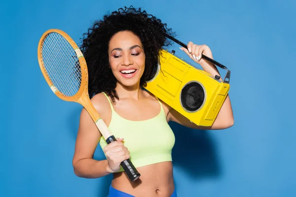Mujer afroamericana feliz sosteniendo boombox y raqueta de tenis en azul - foto de stock