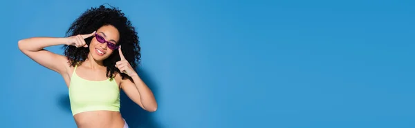 Happy african american woman adjusting sunglasses with fingers on blue, banner — Stock Photo