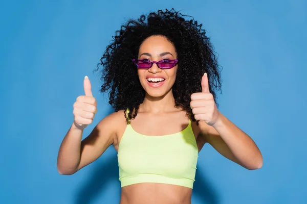 Heureuse afro-américaine dans des lunettes de soleil montrant pouces sur bleu — Photo de stock