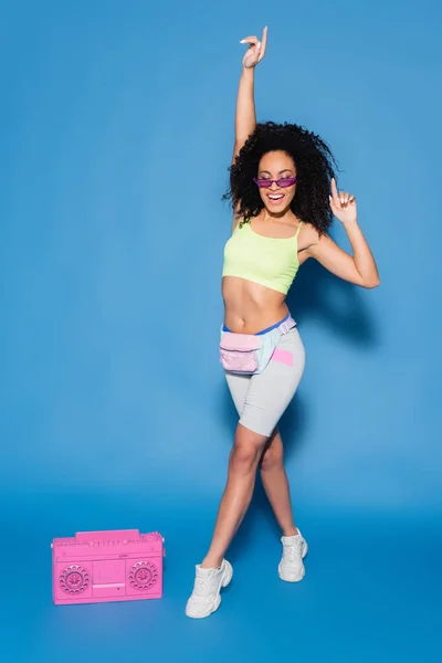 Comprimento total de mulher americana africana sorridente em óculos de sol e bolsa de cintura apontando perto boombox rosa no azul — Fotografia de Stock