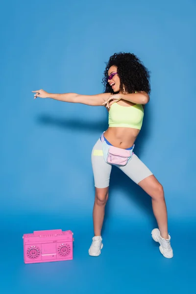 Longitud completa de la mujer afro-americana feliz en gafas de sol y bolsa de cintura apuntando cerca de boombox rosa en azul - foto de stock