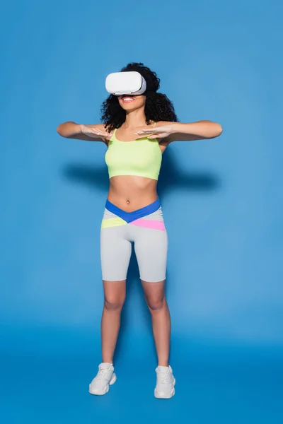 Longitud completa de la mujer afroamericana feliz en auriculares vr ejercicio en azul - foto de stock