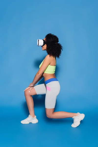 Full length of smiling african american woman in vr headset exercising on blue — Stock Photo