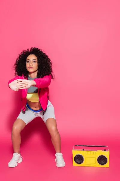 Full length of curly african american woman in jacket exercising near yellow boombox on pink — Stock Photo