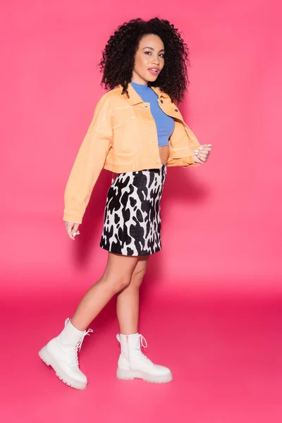 Longitud completa de la mujer afroamericana en falda con estampado animal, chaqueta y top de la cosecha posando en rosa - foto de stock