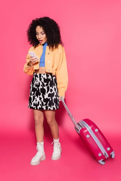 Full length of shocked african american woman using smartphone near suitcase on pink — Stock Photo
