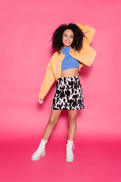 Longitud completa de la mujer afroamericana complacida en falda con estampado animal, chaqueta y top de la cosecha posando en rosa - foto de stock