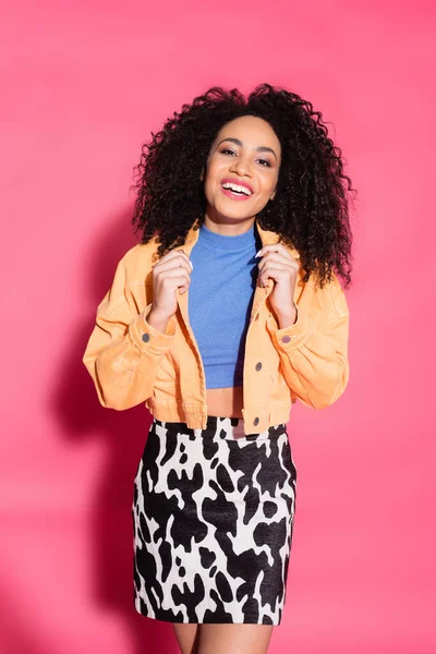 Happy african american woman in skirt with animal print adjusting jacket while posing on pink — Stock Photo