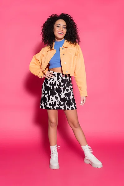 Longitud completa de la mujer afroamericana feliz en falda con estampado de animales, chaqueta y top de la cosecha posando en rosa - foto de stock