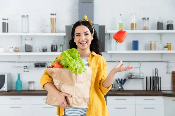 Glückliche brünette Frau wirft Paprika in die Luft und hält Papiertüte mit Lebensmitteln — Stockfoto
