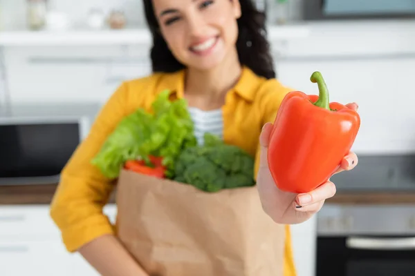 Blurred and happy brunette woman holding paper bag with groceries and bell pepper — Stock Photo