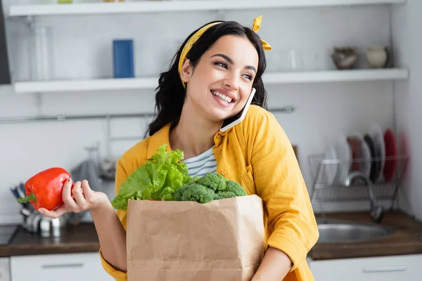 Mujer morena feliz sosteniendo bolsa de papel con comestibles y hablando en el teléfono inteligente - foto de stock