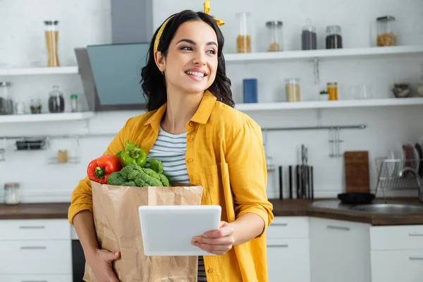 Fröhliche brünette Frau hält Papiertüte mit Lebensmitteln und nutzt digitales Tablet — Stockfoto