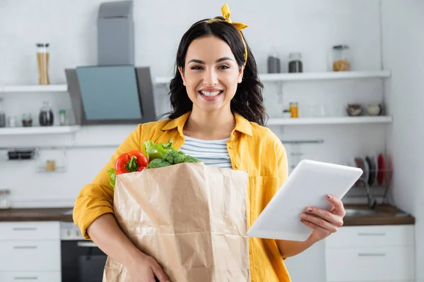 Alegre morena mujer sosteniendo bolsa de papel con comestibles y tableta digital - foto de stock