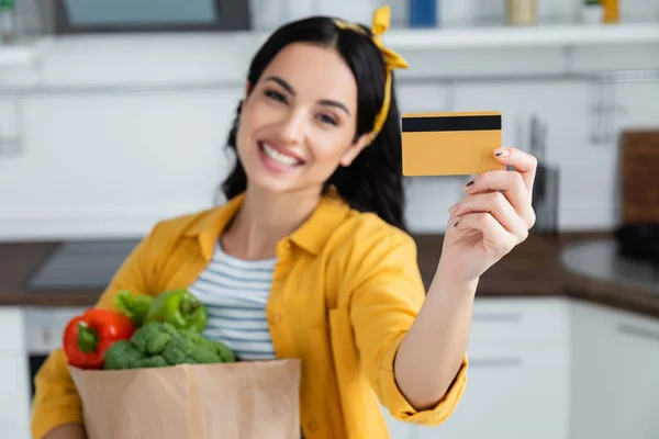 Happy and blurred woman holding paper bag with groceries and credit card — Stock Photo