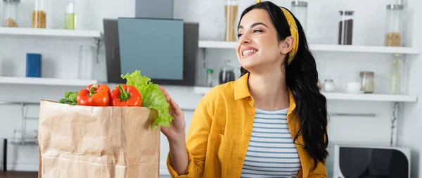 Alegre morena mujer sonriendo cerca de bolsa de papel con verduras, pancarta - foto de stock