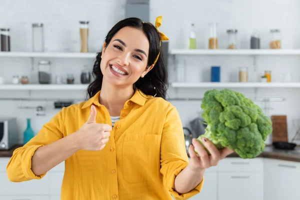 Glückliche brünette Frau hält reifen grünen Brokkoli, während sie den Daumen nach oben zeigt — Stockfoto