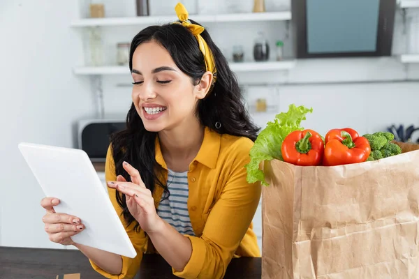 Felice donna bruna utilizzando tablet digitale vicino sacchetto di carta con generi alimentari — Foto stock