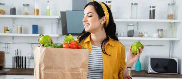 Mujer morena feliz mirando bolsa de papel con alimentos frescos, pancarta - foto de stock