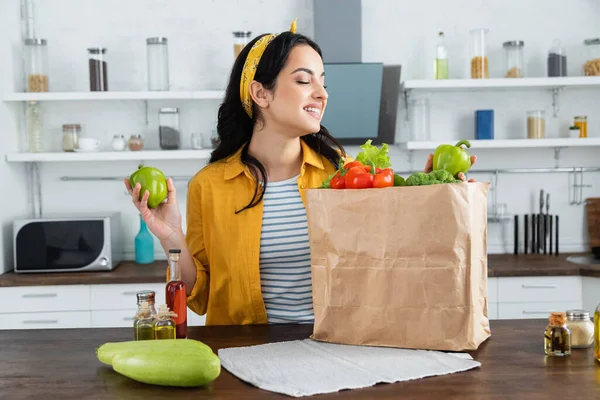 Mujer morena feliz mirando bolsa de papel con alimentos frescos en la mesa de la cocina - foto de stock